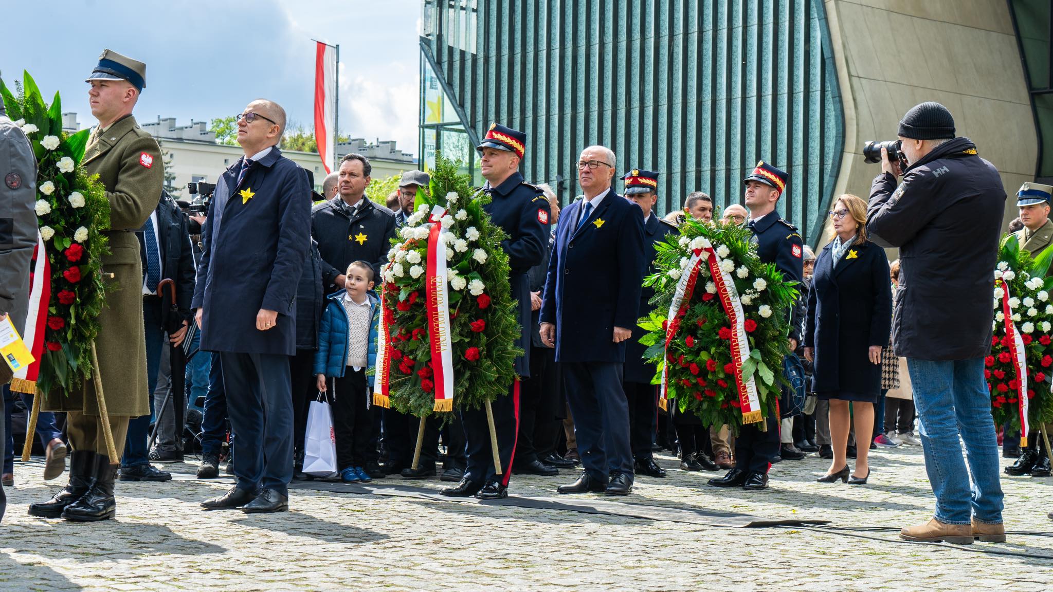 Uczestnicy w mundurach wojskowych i ubraniach cywilnych składają wieńce podczas obchodów 81. rocznicy powstania w getcie warszawskim, przed nowoczesnym budynkiem.