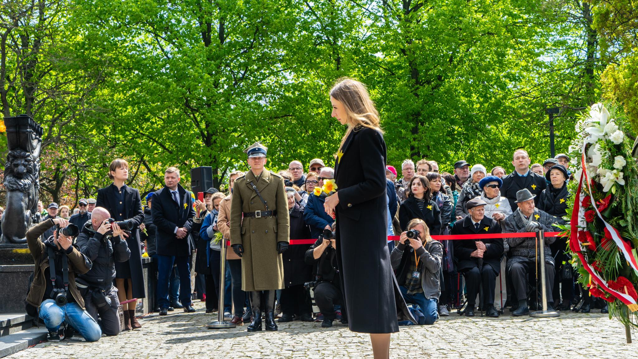 Uroczystość upamiętniająca 81. rocznicę powstania w getcie warszawskim: przed zgromadzonym tłumem stoi kobieta w czarnym płaszu, obok niej oficer w historycznym mundurze, a fotografowie na kolanach robią zdjęcia obok dużego wieńca.