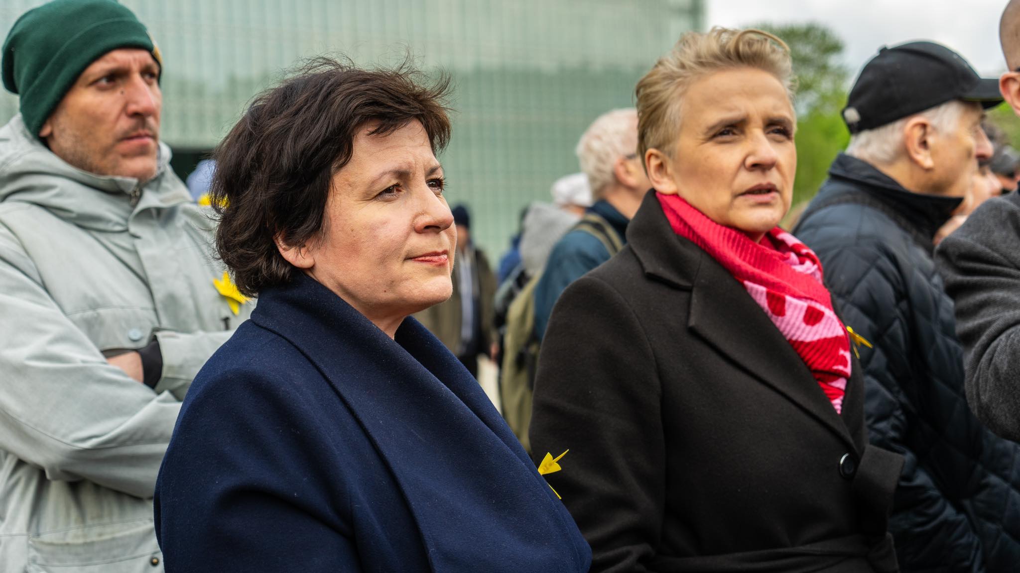 Grupa uczestników obchodów 81. rocznicy powstania w getcie warszawskim; na pierwszym planie dwie kobiety w ciemnych płaszczach z żółtymi wstążkami przypiętymi do ubrań.