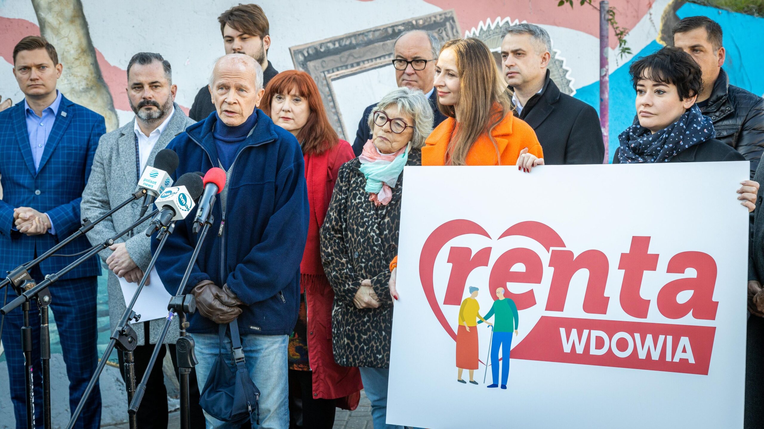 Na zdjęciu widoczna jest grupa osób stojących przed plakatem promującym rentę wdowią, z mówcą trzymającym mikrofon na pierwszym planie.