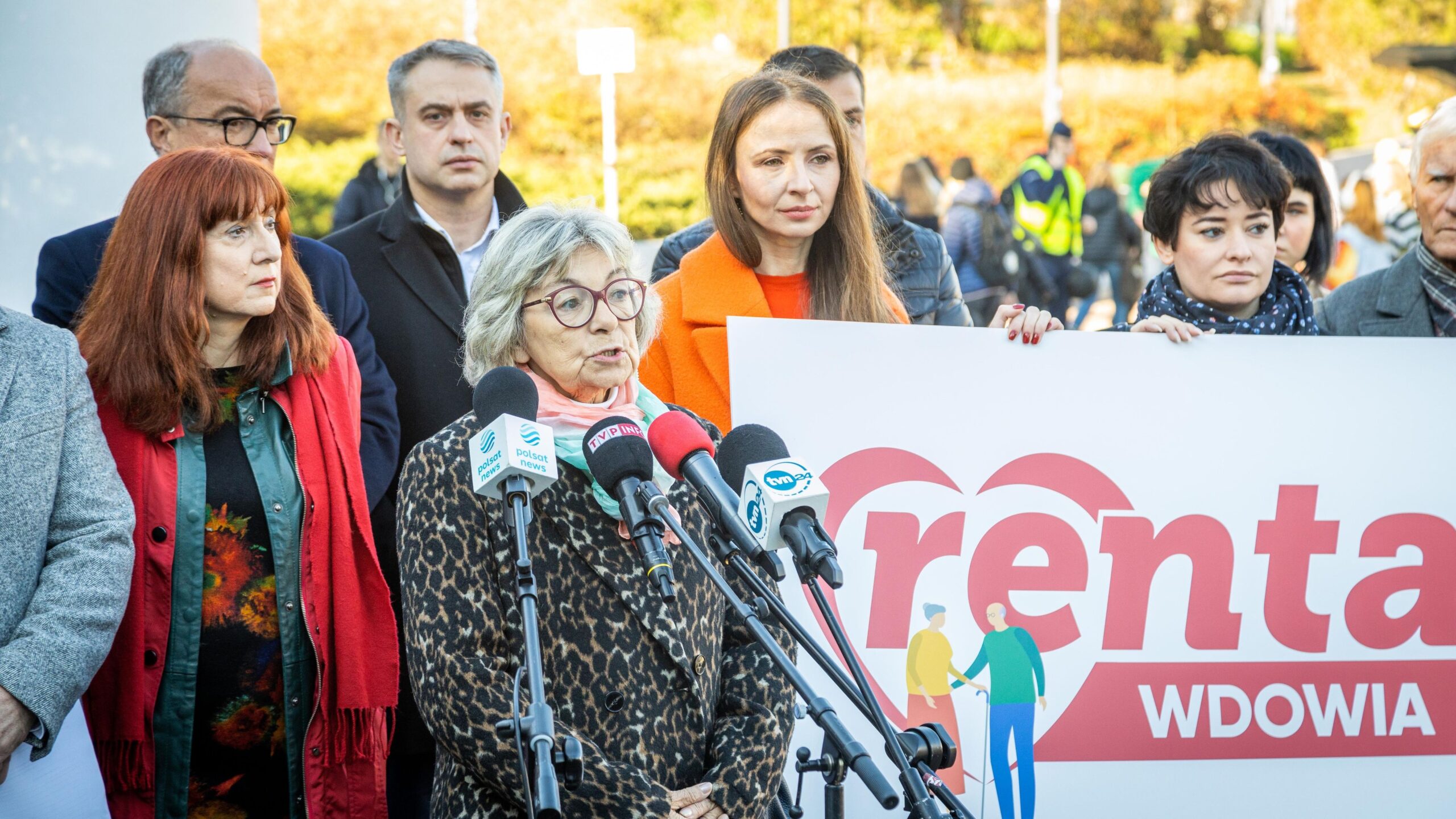 Na zdjęciu przedstawiono grupę ludzi, w tym kobietę przemawiającą przy mikrofonach podczas wydarzenia związanego z kampanią publiczną dotyczącą renty wdowiej, z widocznym dużym banerem z napisem 