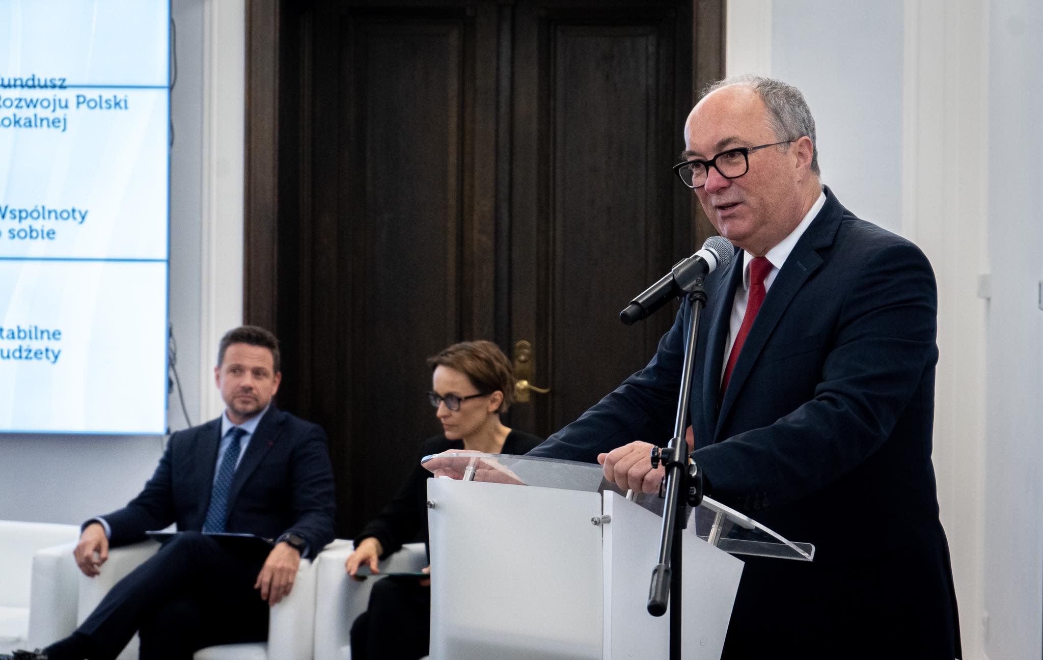 Polityk w garniturze i czerwonym krawacie przemawia z mikrofonem przy mównicy, podczas gdy dwie osoby siedzą na białych fotelach w tle.