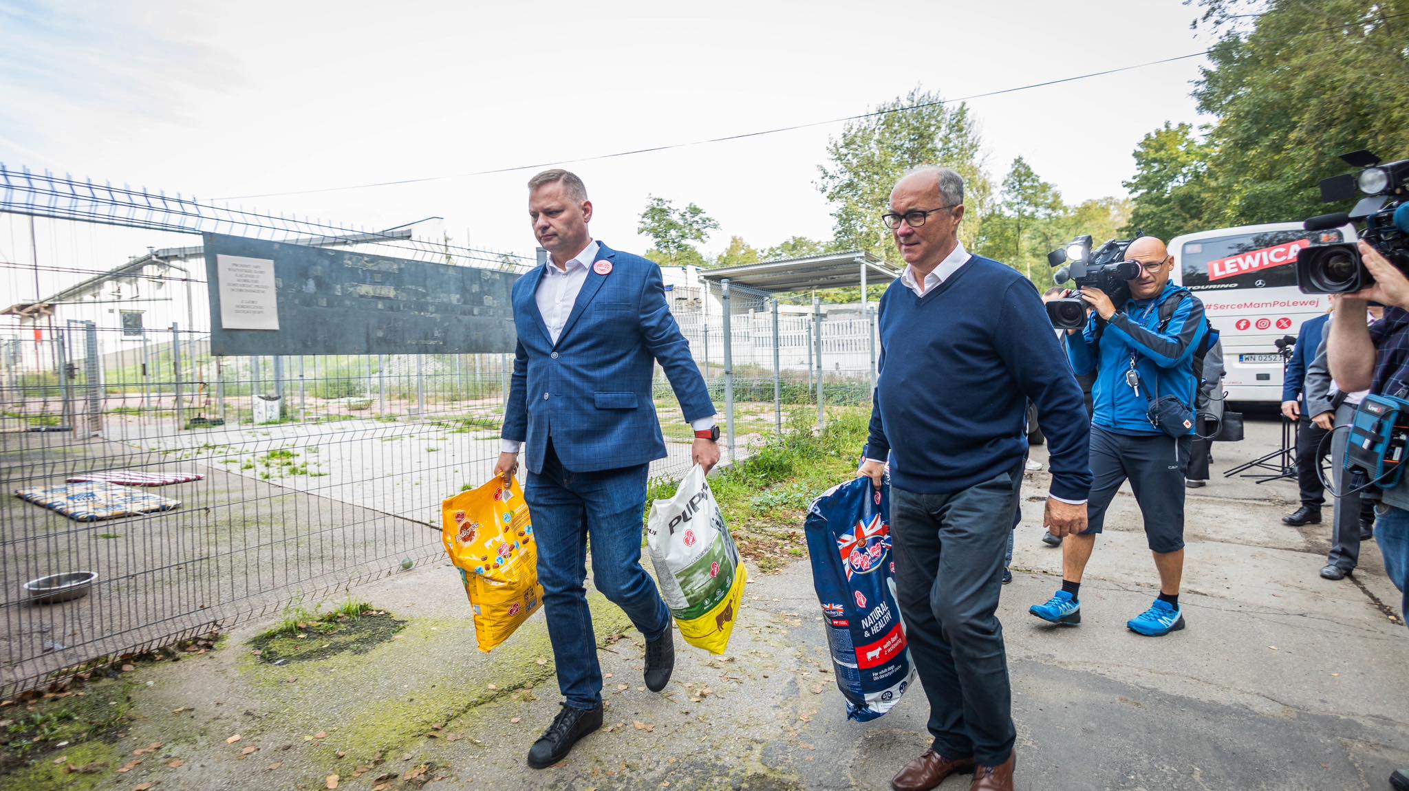 Dwóch mężczyzn niesie worki z karmą dla zwierząt podczas wizyty w schronisku, a w tle pracują ekipy medialne z kamerami i autobus z napisem Lewica.