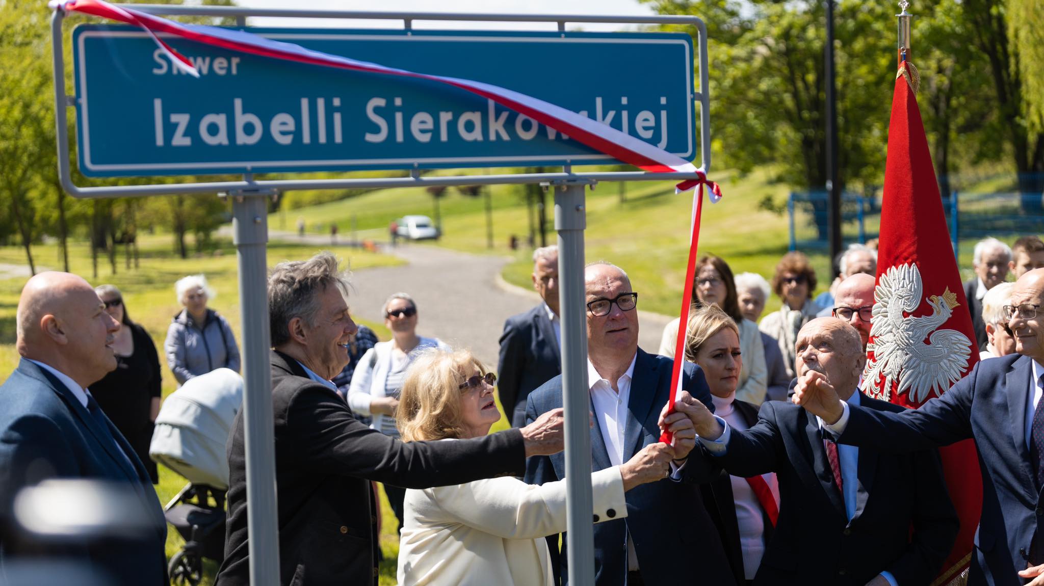 Uroczystość odsłonięcia tablicy z nazwą skweru Izabelli Sierakowskiej, w której uczestniczy grono osób w różnych strojach oraz flagą Polski w tle.