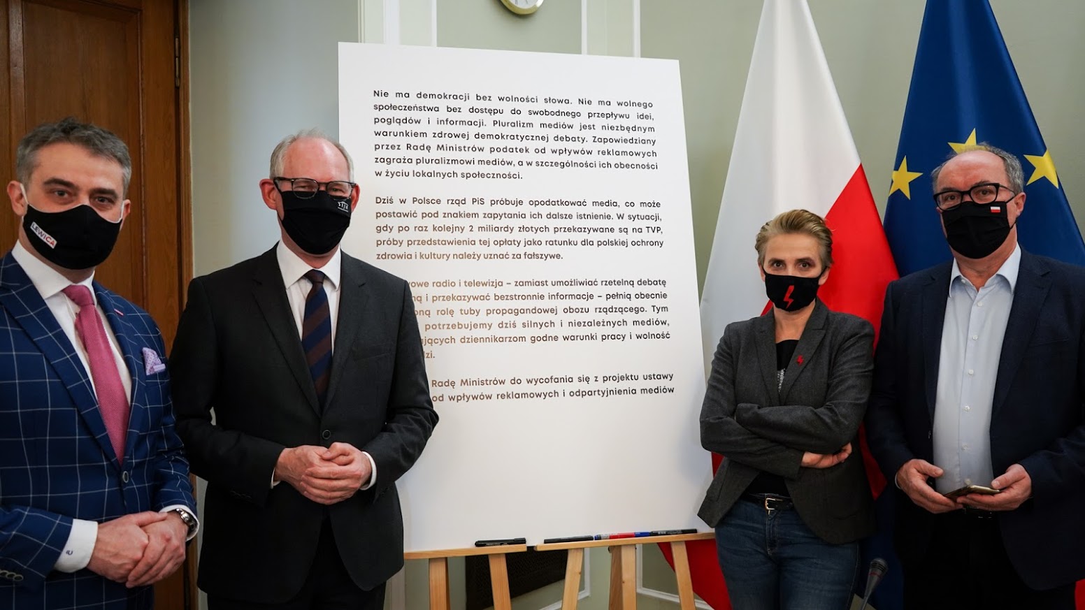 Czterech osób w maskach i eleganckich ubraniach stoi obok tablicy z tekstem, na tle flag Polski i Unii Europejskiej, podczas spotkania dotyczącego odrzucenia podatku od reklam.