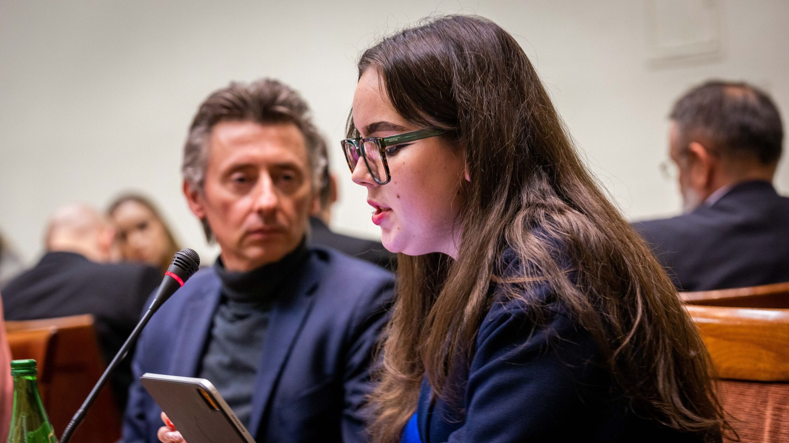 Młoda kobieta z długimi włosami i okularami przemawia do mikrofonu podczas spotkania Okrągły Stół o problemach w psychiatrii dziecięcej.