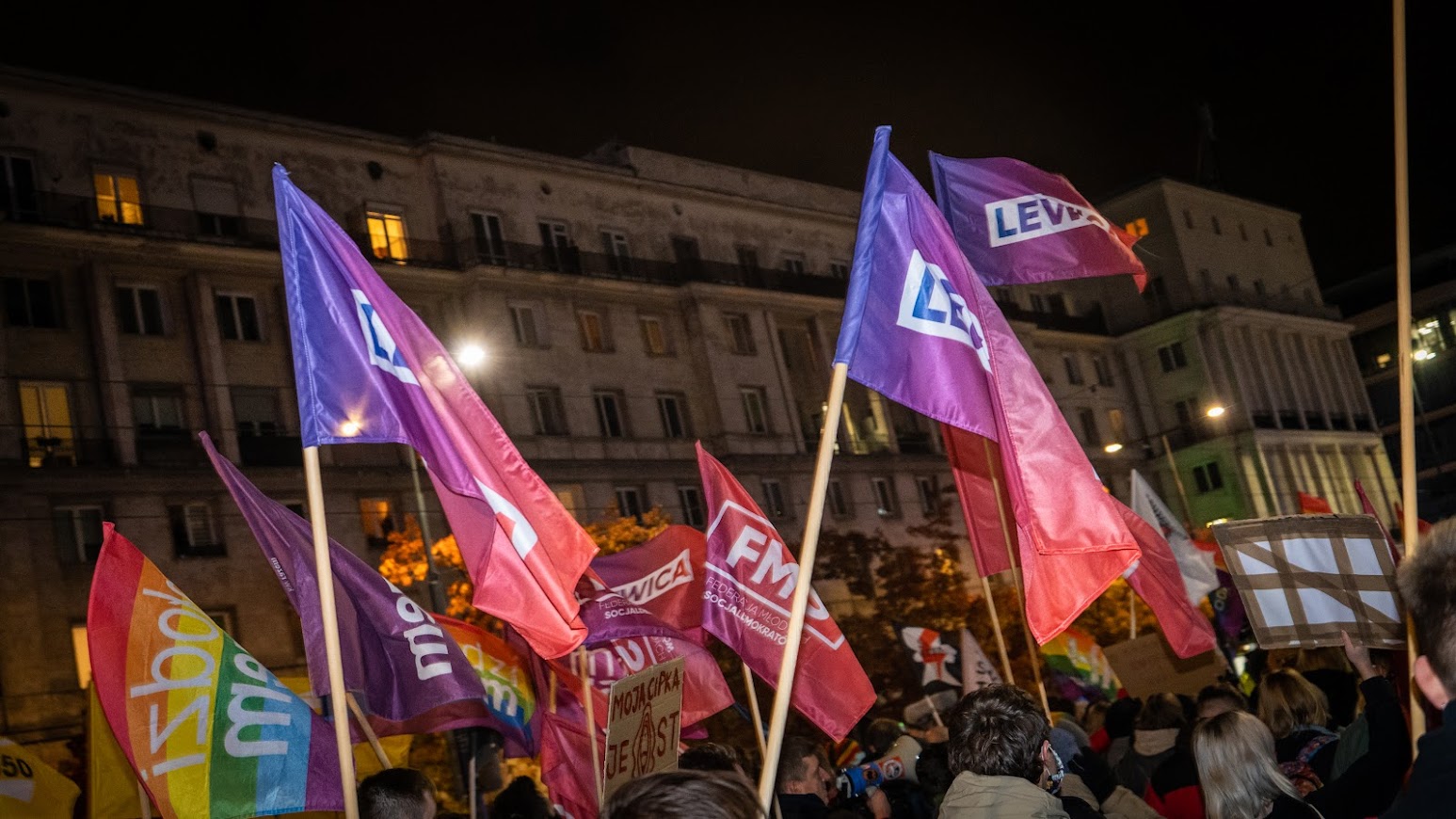 Na zdjęciu widać tłum ludzi niosących kolorowe flagi w nocy podczas manifestacji.