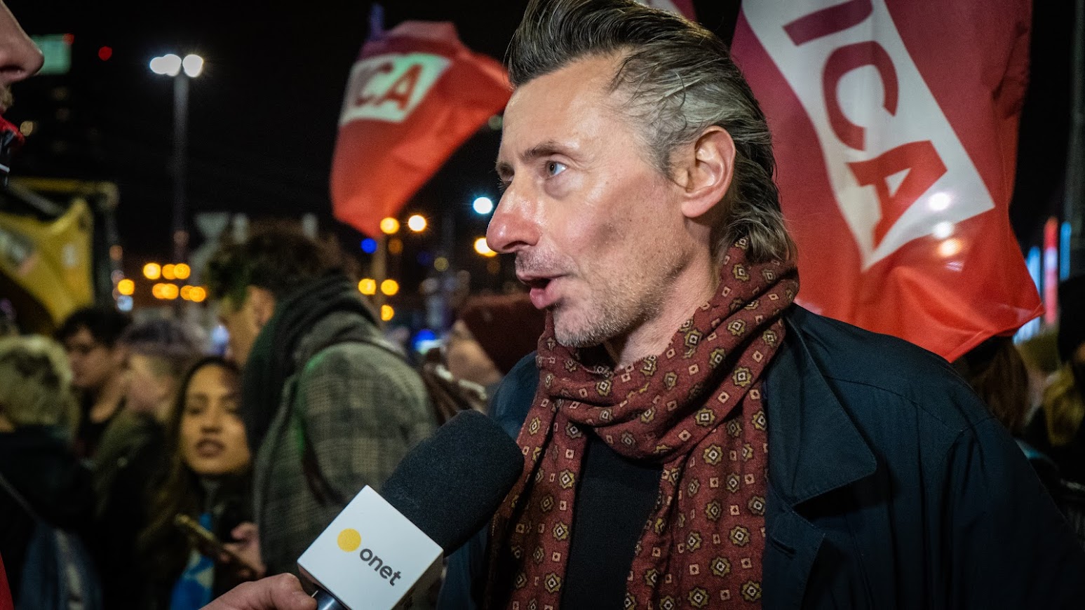 Mężczyzna w szaliku z mikrofonem w ręku rozmawia podczas protestu, a w tle widać uczestników z flagami.