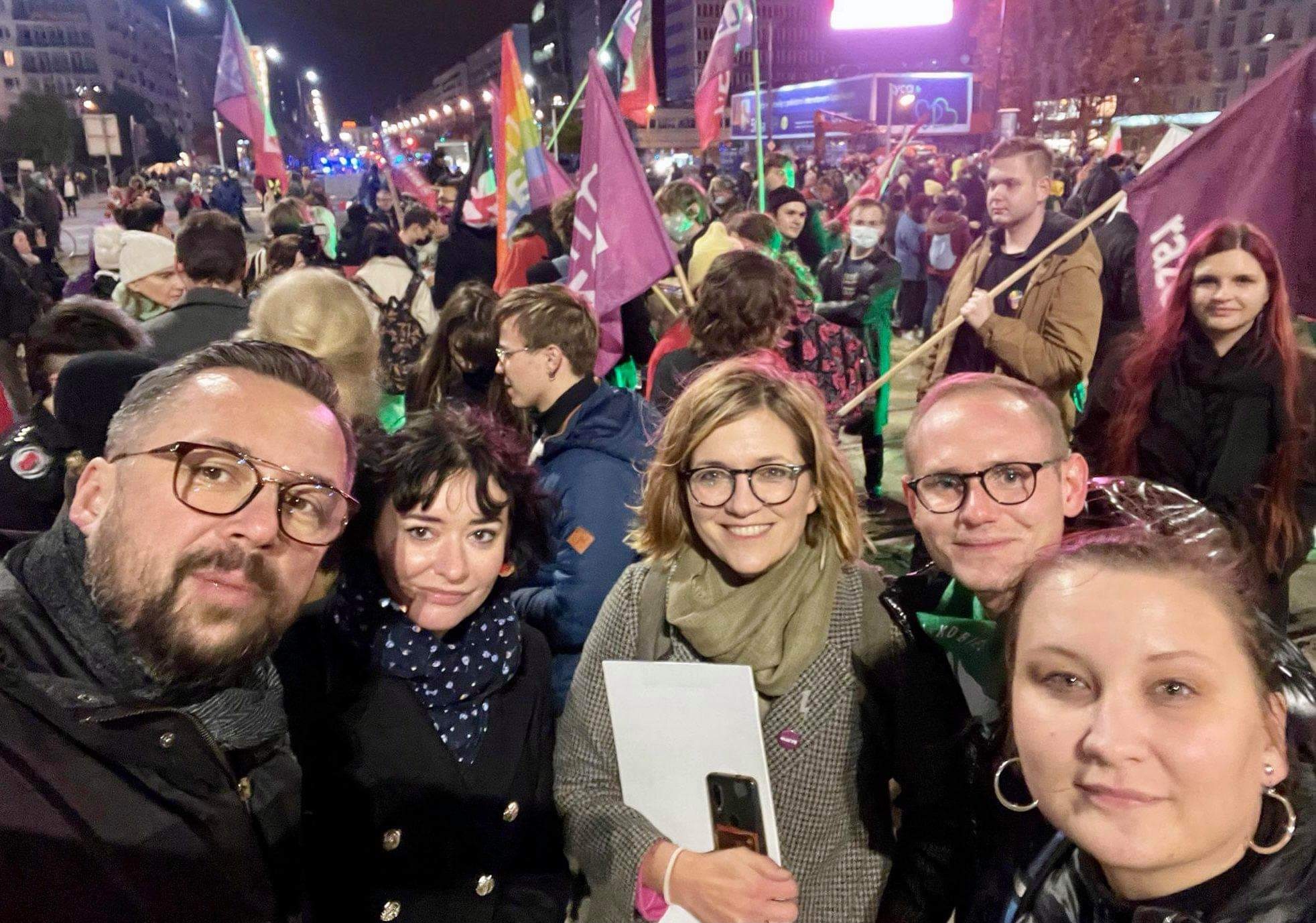 Grupa osób pozujących do zdjęcia podczas protestu, w tle widoczni demonstranci z flagami i oświetleniem miejskim w nocy.