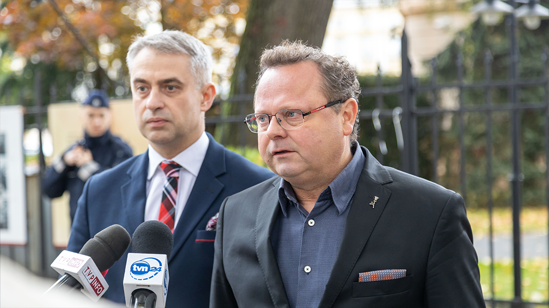 Dwaj mężczyźni w garniturach występują przed mikrofonami, otoczeni jesiennym tłem z drzewami.