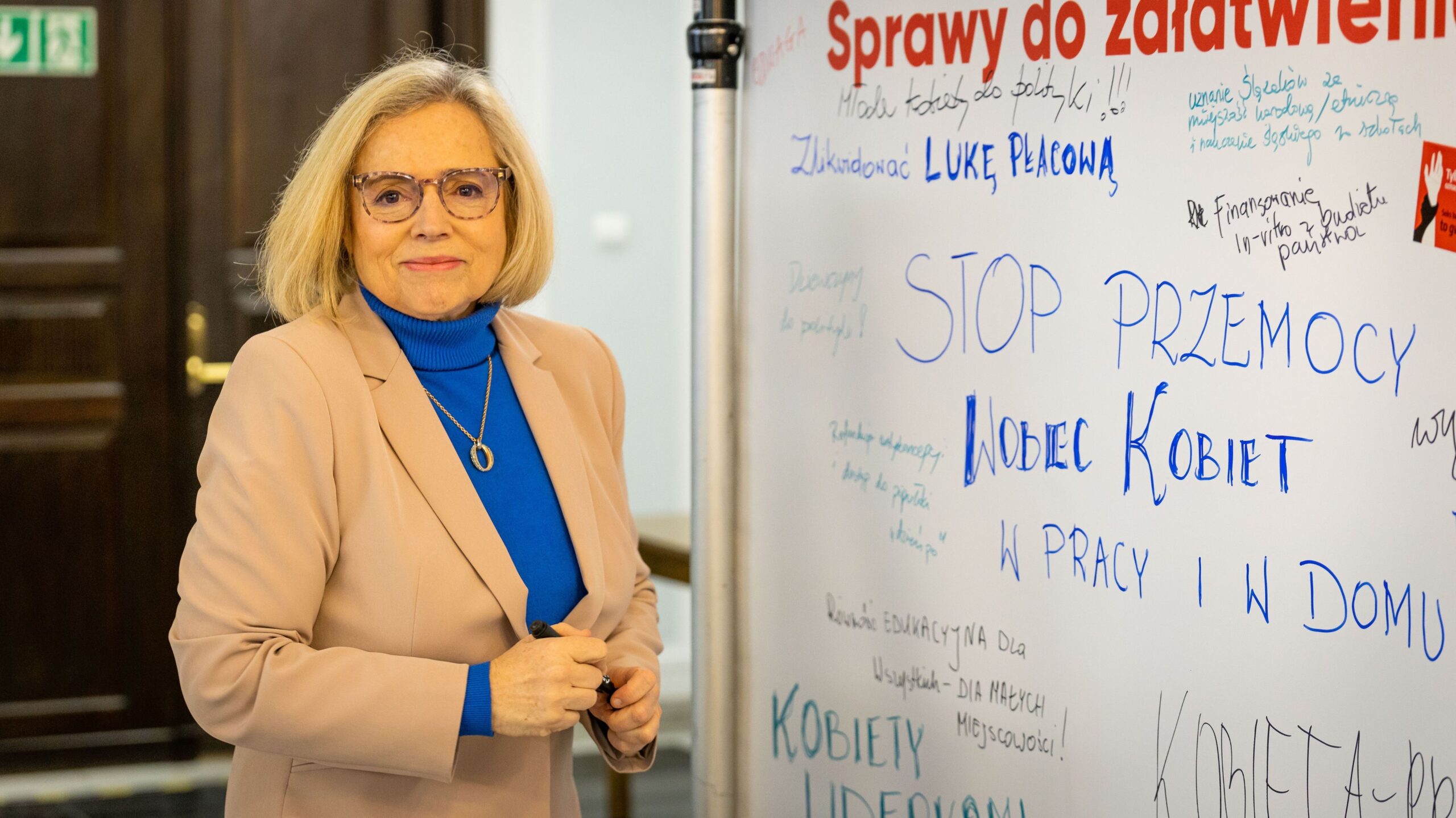 Na zdjęciu widoczna jest kobieta w beżowym żakiecie, która stoi obok tablicy z hasłami dotyczącymi praw kobiet.