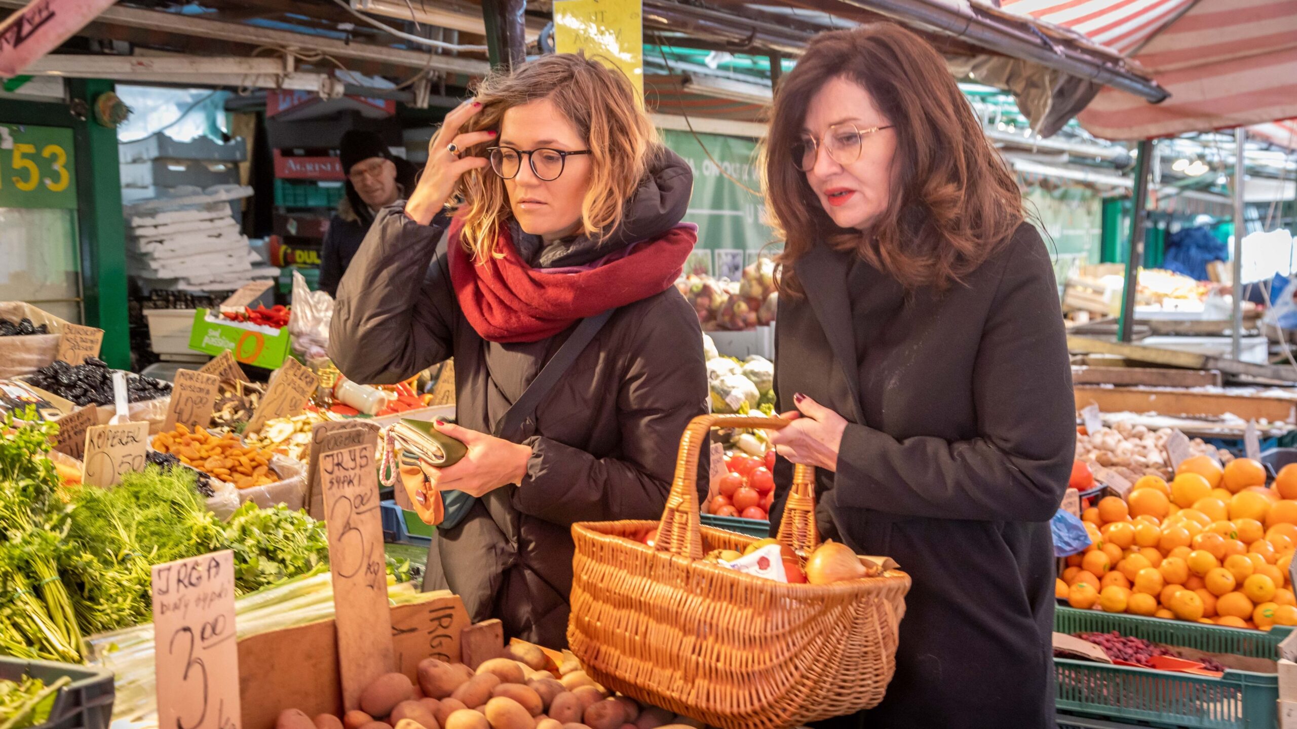 Na zdjęciu widać dwie kobiety, które zastanawiają się nad zakupem produktów na targowisku, w otoczeniu owoców i warzyw.