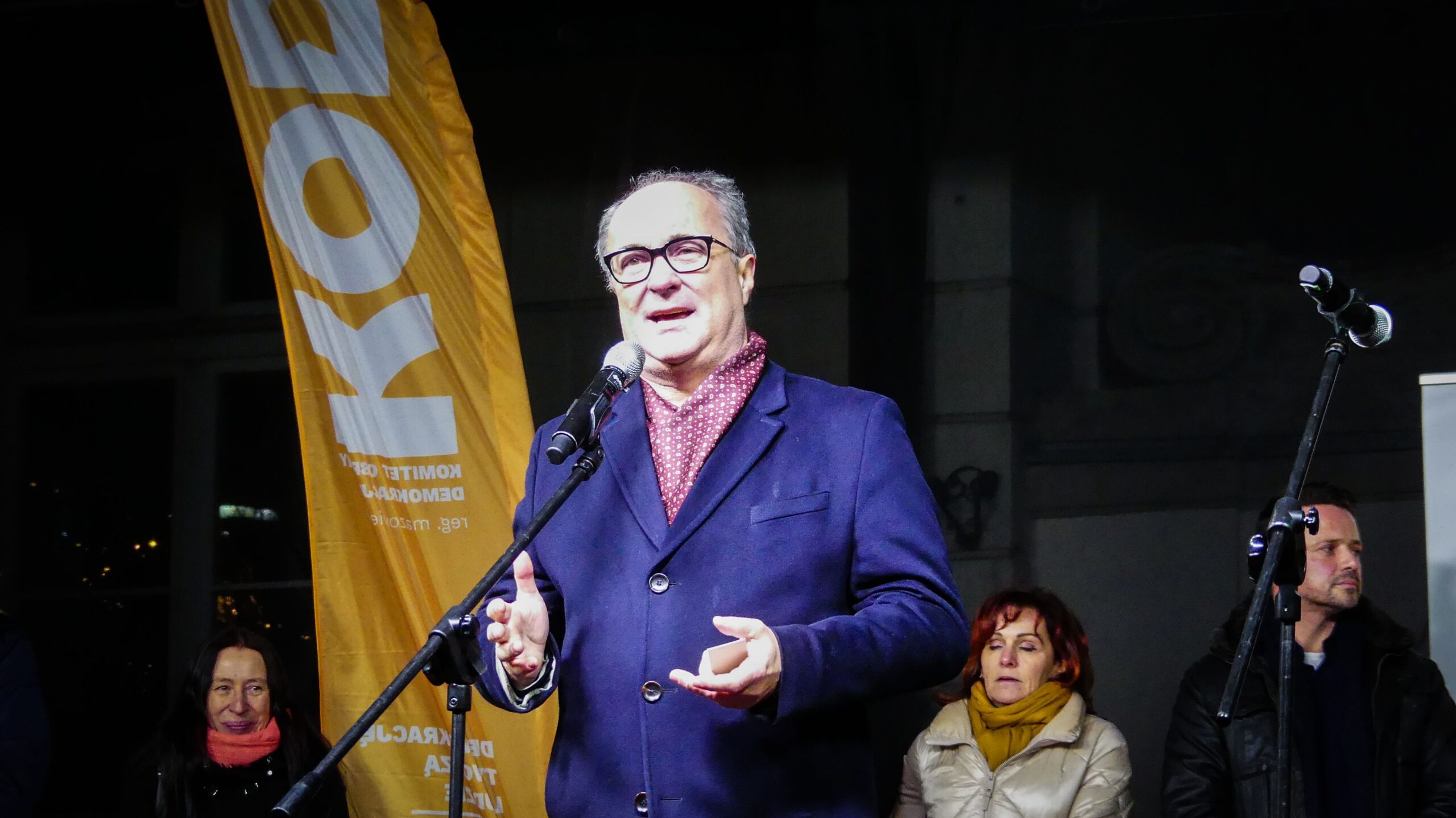 Na zdjęciu widoczny jest mężczyzna mówiący do mikrofonu na tle żółtej flagi, a w tle siedzą osoby słuchające jego wystąpienia.