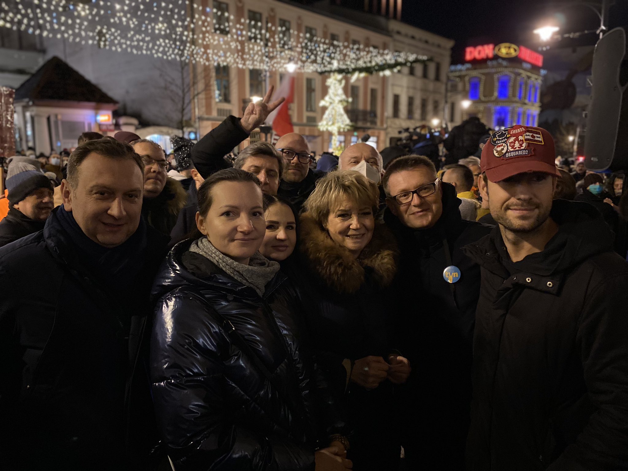 Grupa ludzi w zimowych kurtkach robi wspólne zdjęcie na nocnym zgromadzeniu „Nie dla Lex TVN”, na tle miejskiego placu z choinką i świątecznymi lampkami.