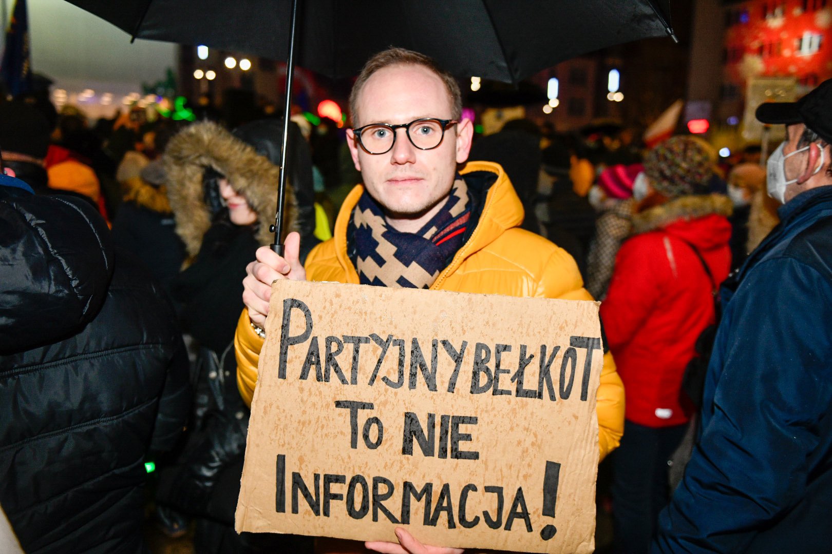 Na zdjęciu widoczny jest mężczyzna w żółtej kurtce trzymający transparent z napisem „Partyjny bełkot to nie informacja!”, na tle tłumu podczas protestu.