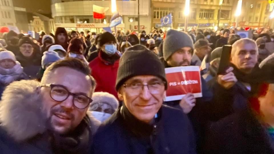 Dwóch mężczyzn na pierwszym planie robi selfie podczas wieczornego protestu „Nie dla Lex TVN”, w tłumie widać plakat i flagi.