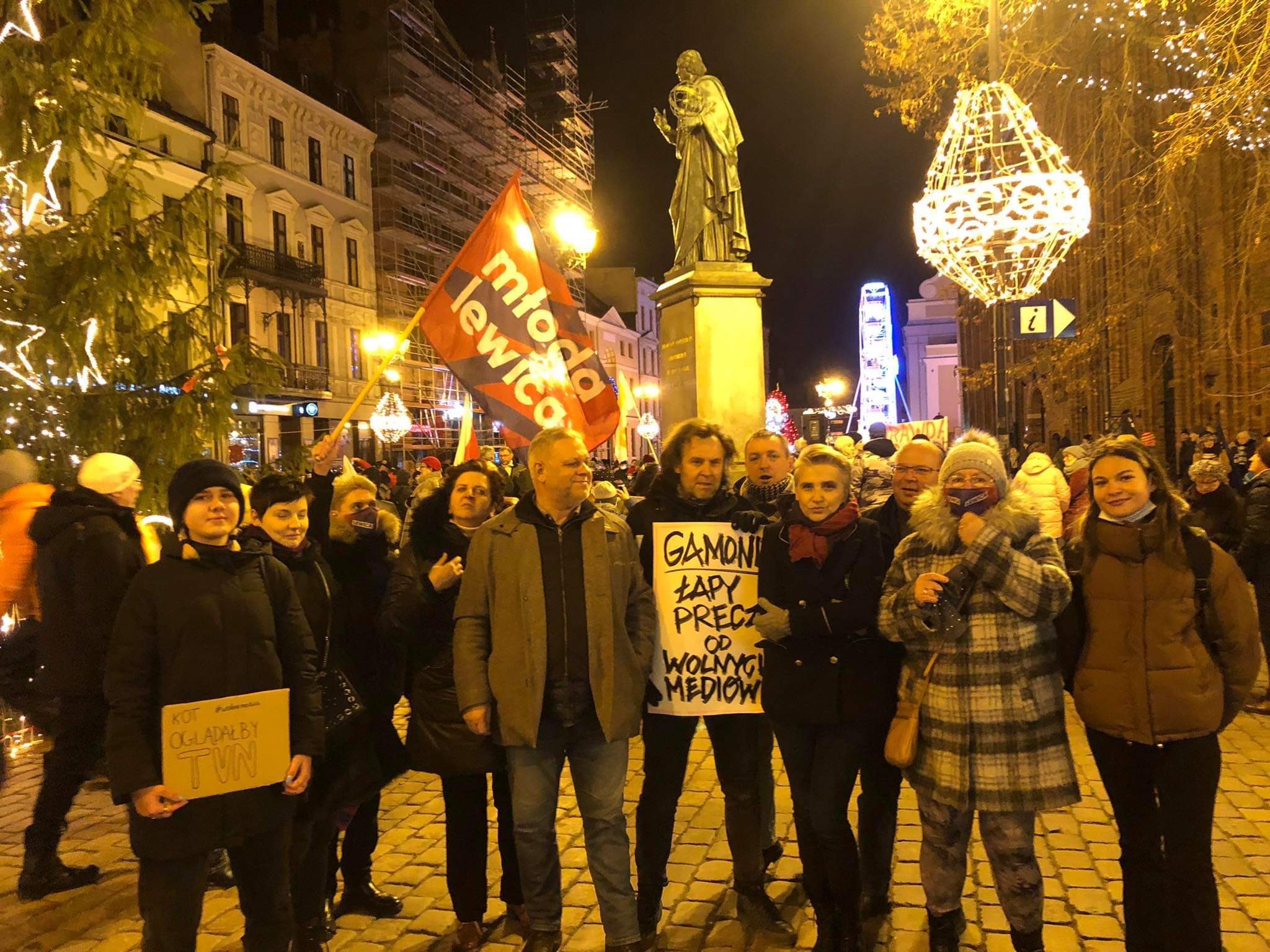 Grupa demonstrantów na miejskim placu wieczorem, trzymająca plakaty i czerwono-białą flagę z napisem „Młoda Lewica”, w tle posąg i świąteczne iluminacje.
