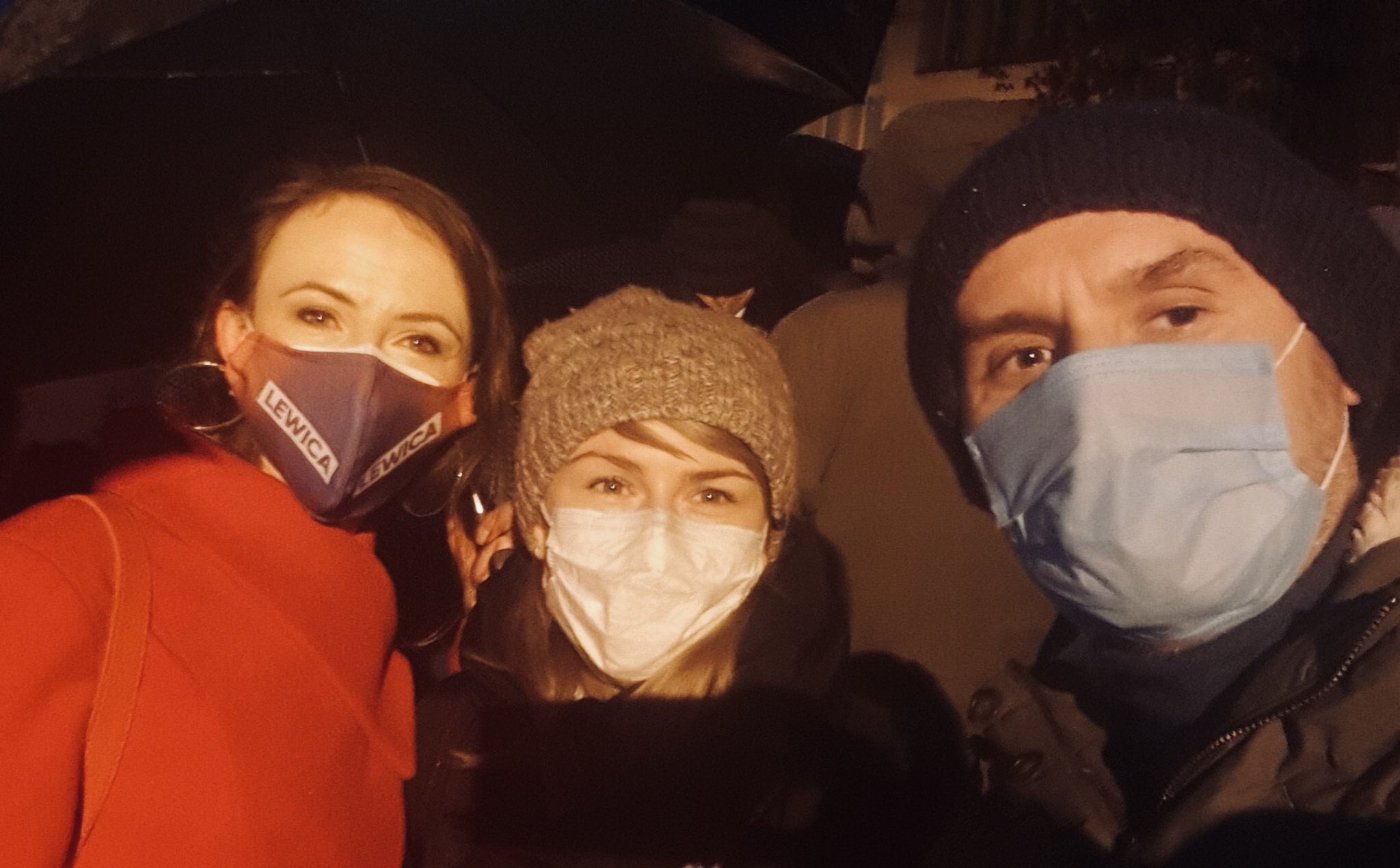 Trzy osoby na zewnątrz wieczorem, wszyscy w maseczkach, robią sobie wspólne zdjęcie; po lewej kobieta w pomarańczowym płaszczu z maską z napisem LEWICA, na środku kobieta w szarej czapce i białej masce, po prawej mężczyzna w czapce i niebieskiej maseczce.