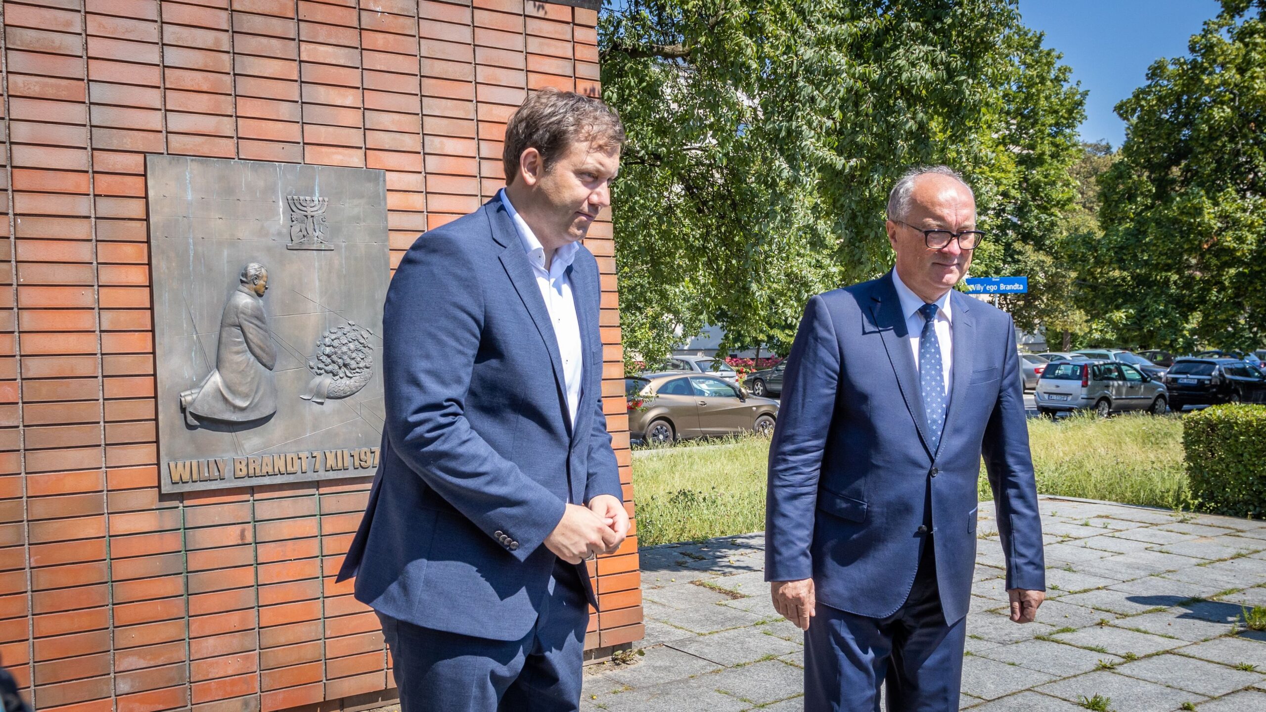 Na zdjęciu widać dwóch mężczyzn w garniturach spacerujących obok tablicy upamiętniającej Willy'ego Brandta.