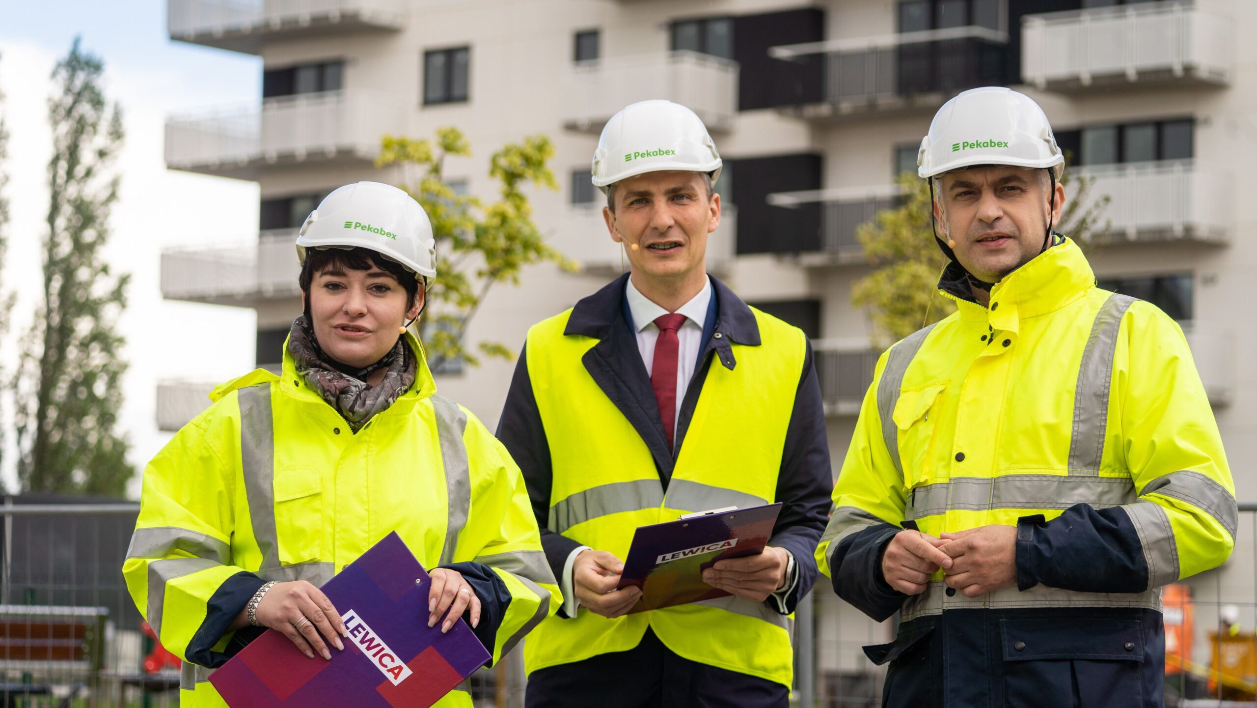 Trójka pracowników budowy w kaskach i żółtej odzieży ochronnej stoi przed nowoczesnym blokiem mieszkalnym; po środku stoi mężczyzna w garniturze trzymający teczkę, po lewej kobieta z fioletowym folderem z napisem Lewica, a po prawej mężczyzna.