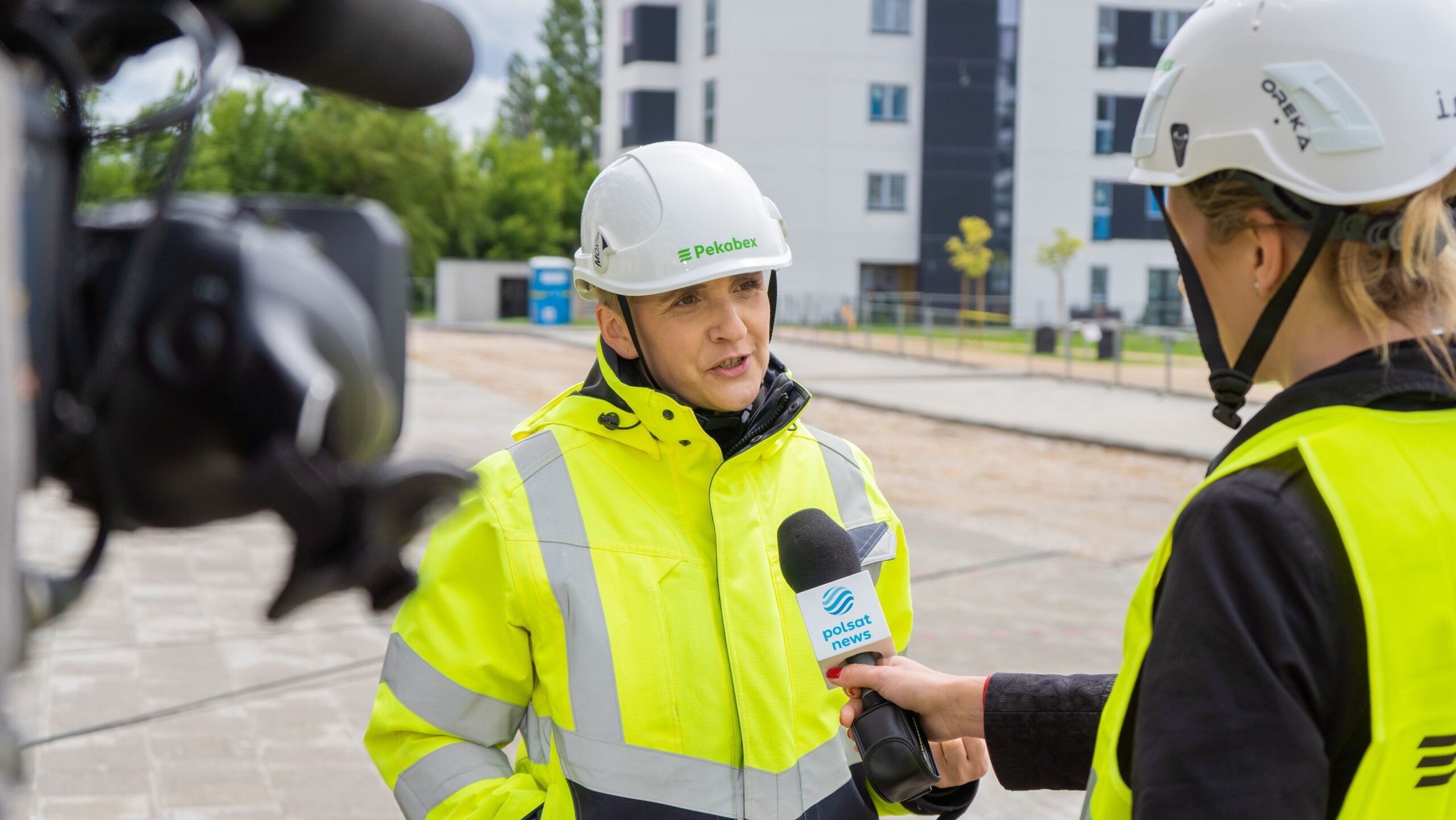 Na zdjęciu widoczna jest osoba w żółtym odblaskowym stroju roboczym, która udziela wywiadu dziennikarce trzymającej mikrofon, a w tle widać budowę.