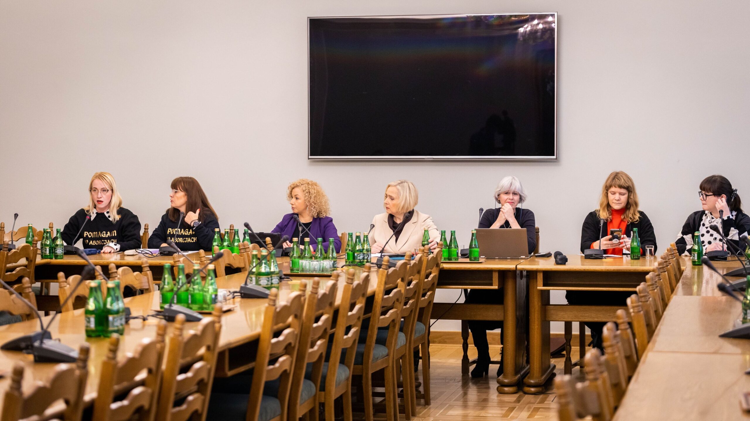 Grupa kobiet siedzi przy długim stole w sali konferencyjnej, niektóre w czarnych koszulkach z napisem, a na stole oraz wokół niego stoi wiele zielonych butelek wody, zaś na ścianie wisi duży monitor.