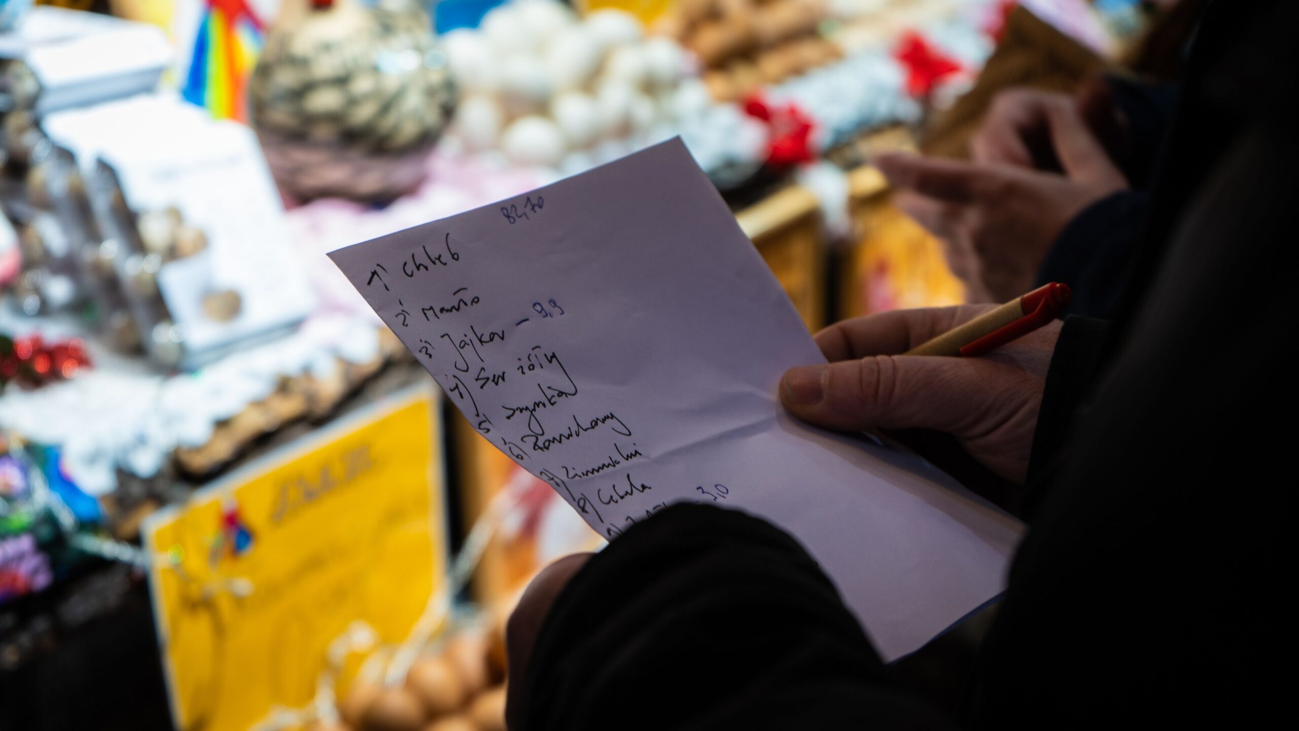 Na zdjęciu widoczna jest osoba trzymająca kartkę z zakupami, na tle kolorowych stoisk z różnymi towarami.