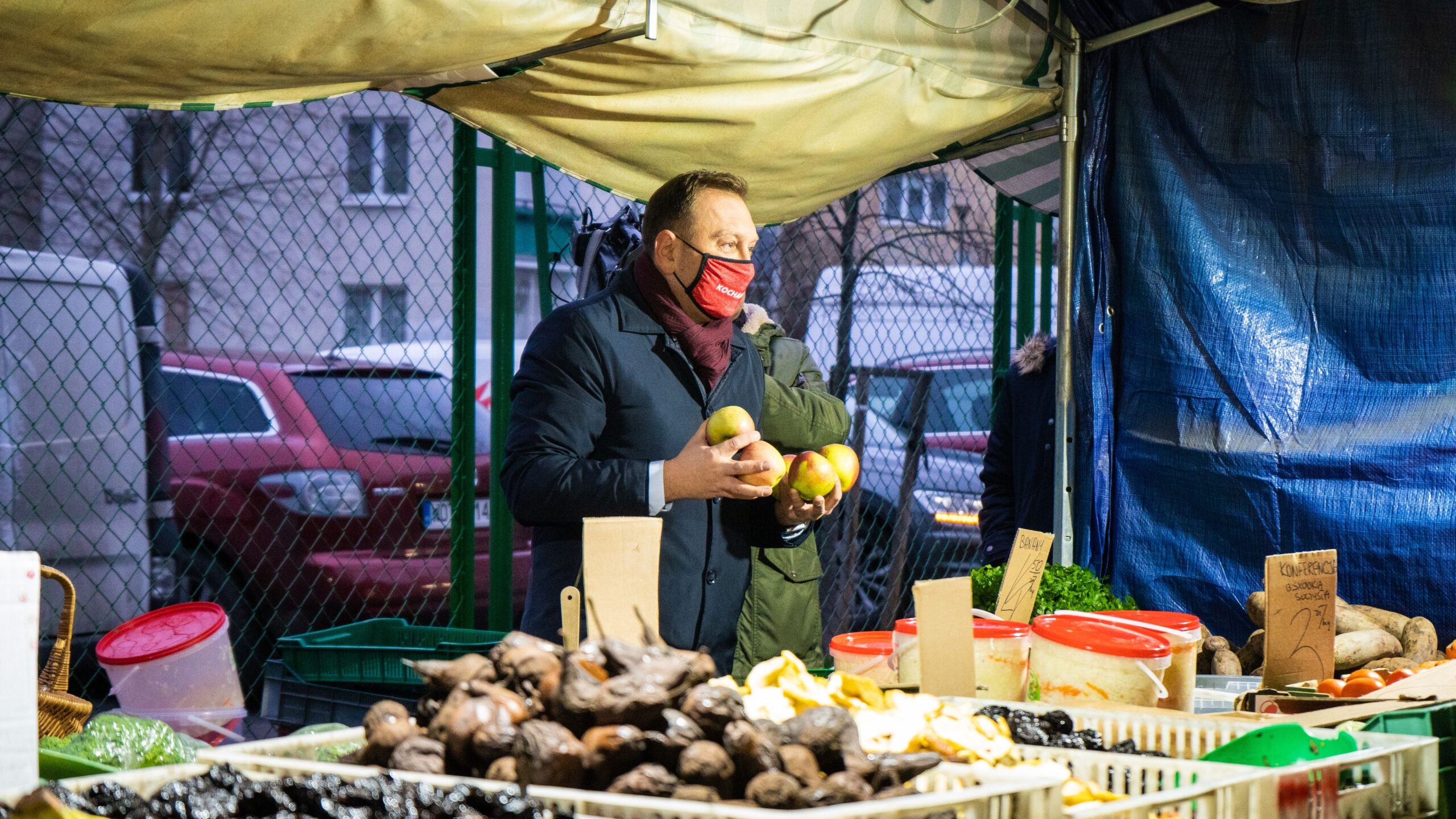 Na zdjęciu mężczyzna w masce trzyma kilka jabłek, przebywając na targu z warzywami i owocami.