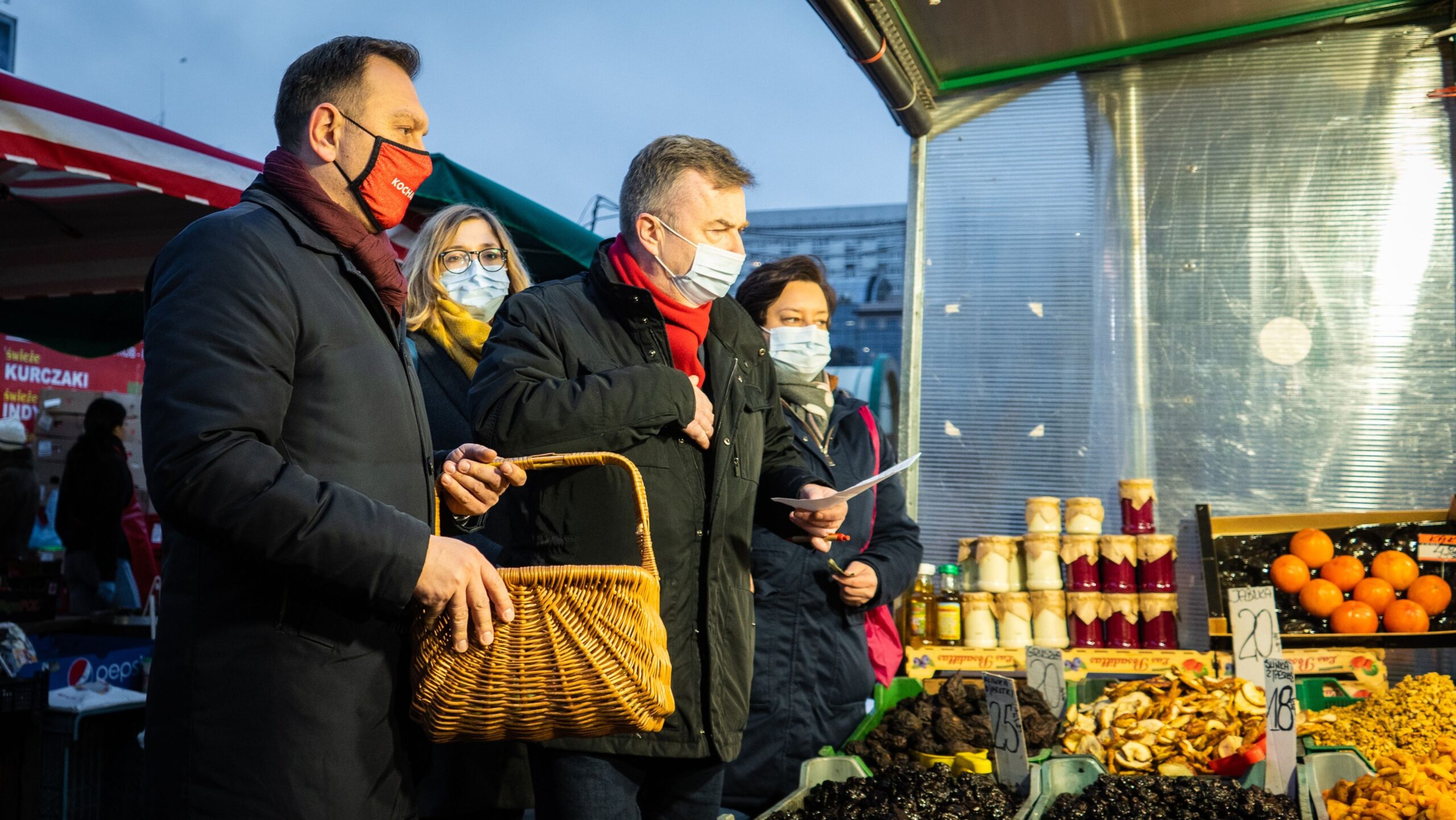 Na zdjęciu cztery osoby w maseczkach stoją przy straganie na rynku, z koszykiem i kartką w rękach, wśród warzyw i przetworów.