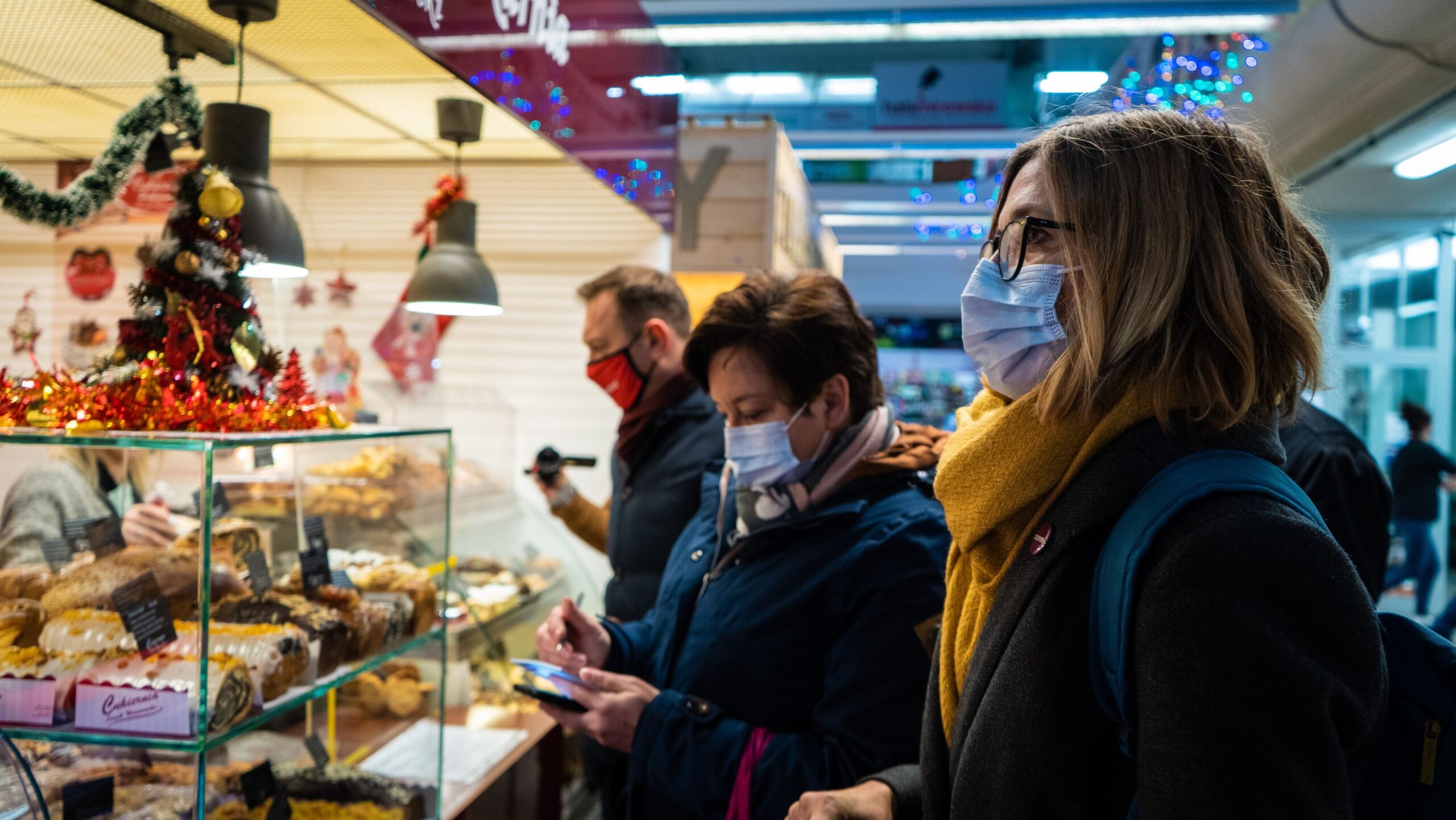 Na zdjęciu widać grupę osób w maseczkach, które przeglądają wypieki w kawiarni ozdobionej świątecznymi dekoracjami.