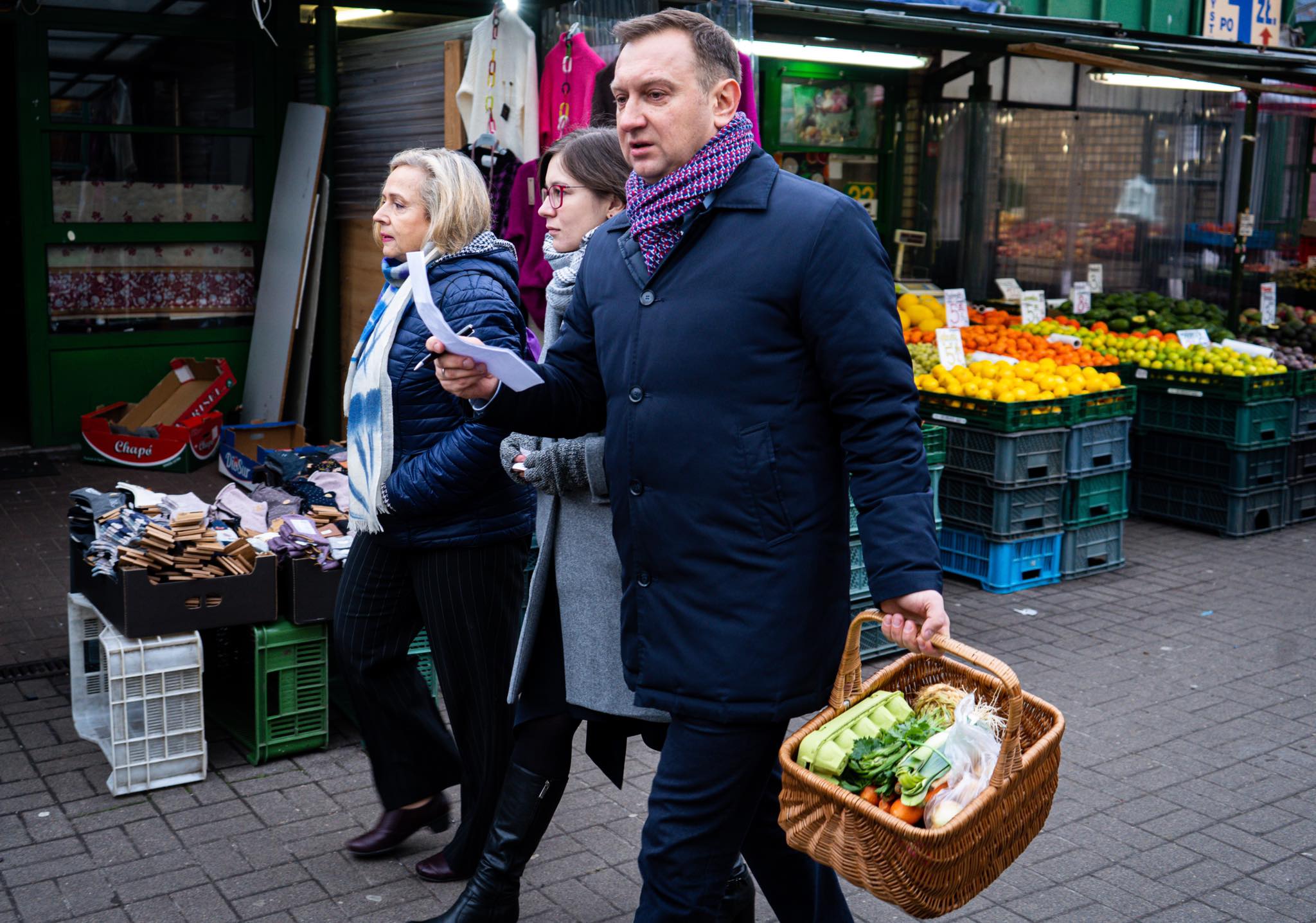 Trzy osoby spacerują po targu, jedna z nich niesie kosz z warzywami, a za nimi widać stragany z owocami i innymi produktami.
