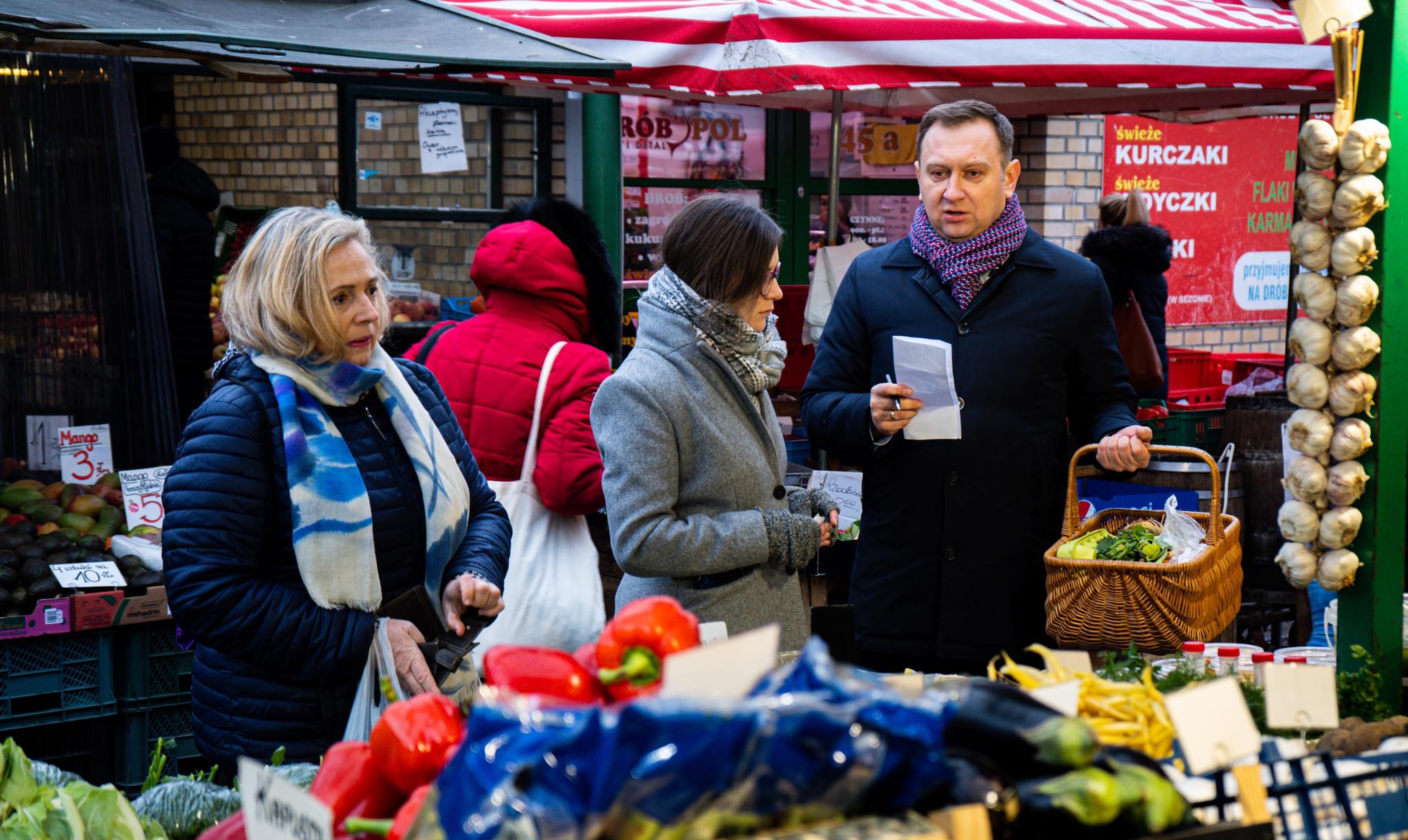 Na zdjęciu widać trzy osoby na targu, które rozmawiają i przeglądają świeże warzywa oraz owoce w koszyku.