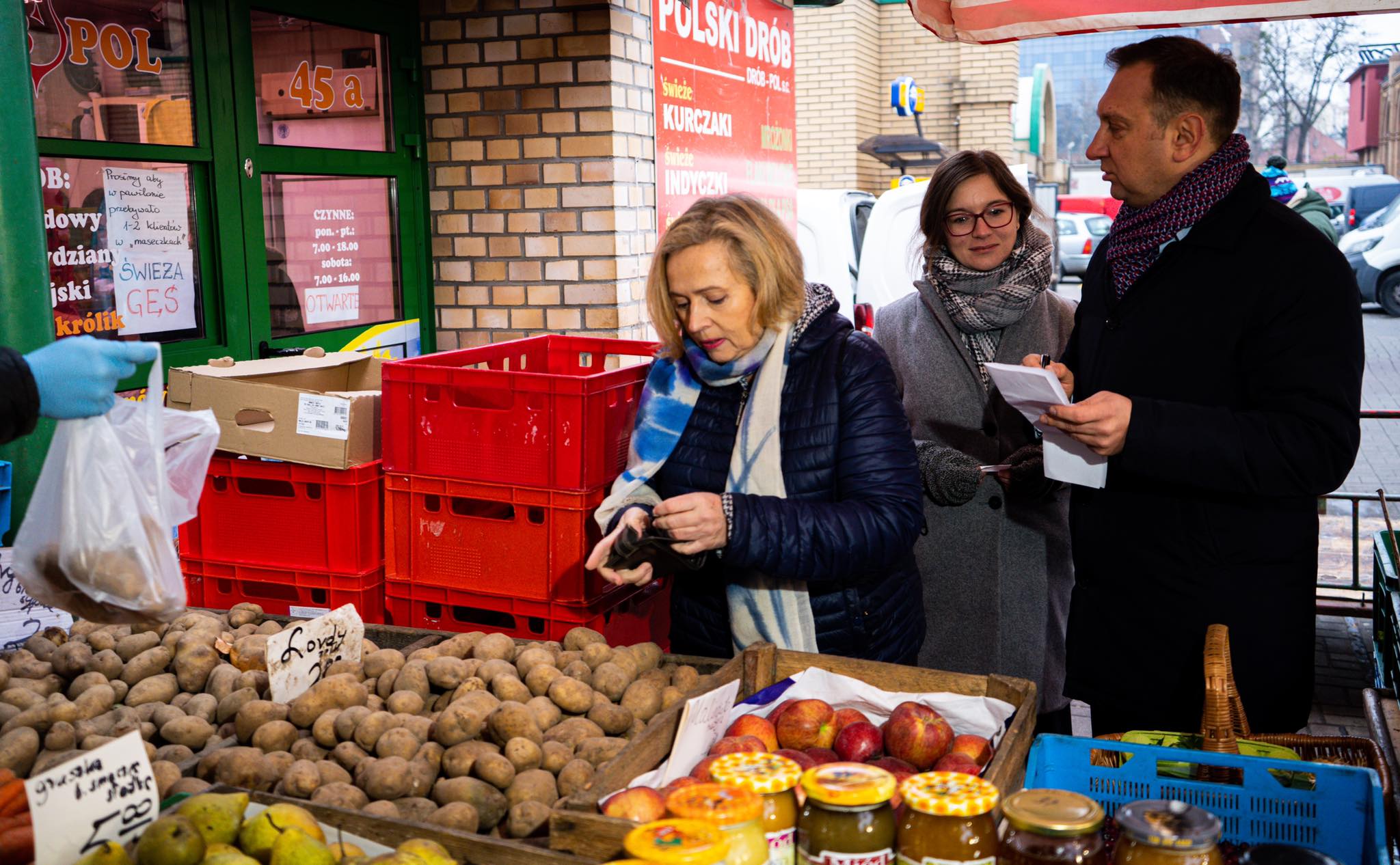 Na zdjęciu widać trzy osoby dokonujące zakupów na lokalnym targu, przy stoisku z ziemniakami i innymi produktami spożywczymi.