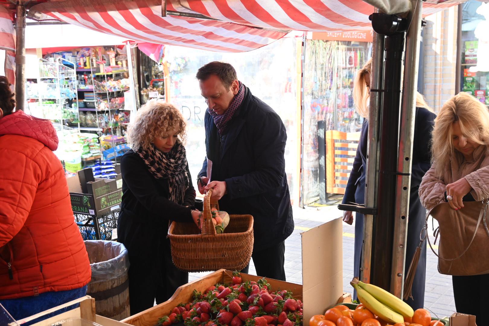 Para na targu owocowo-warzywnym wybiera świeże owoce z kosza, a na stoisku widać skrzynki z truskawkami i bananami.